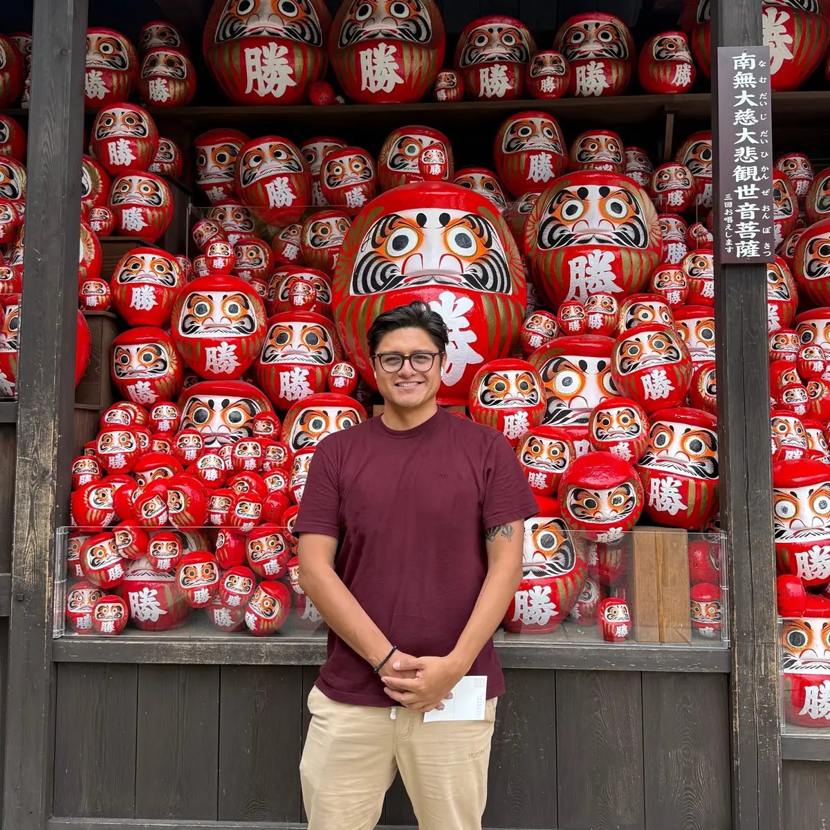 Carlos standing in front of wooden shelves packed with red daruma dolls, wearing a maroon t-shirt, beige trousers and dark-framed glasses, hands clasped, smiling